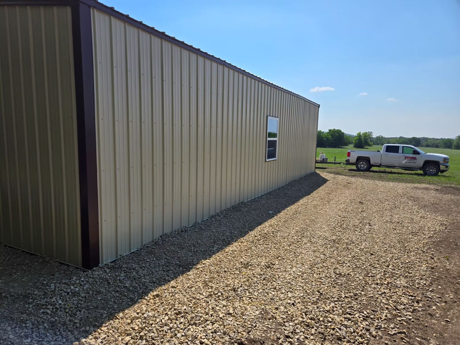 Tan metal building with brown trim, small window, and gravel pathway. A white truck is parked nearby on a sunny day.
