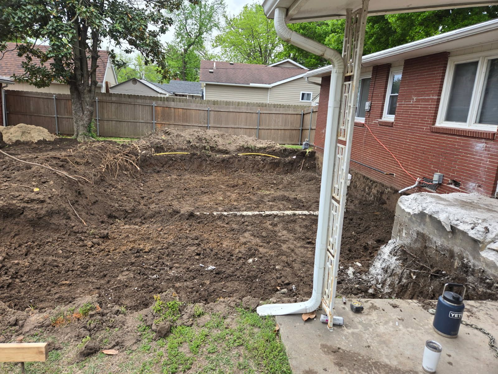 Backyard with freshly dug soil, wooden fence, and a brick house patio in the foreground