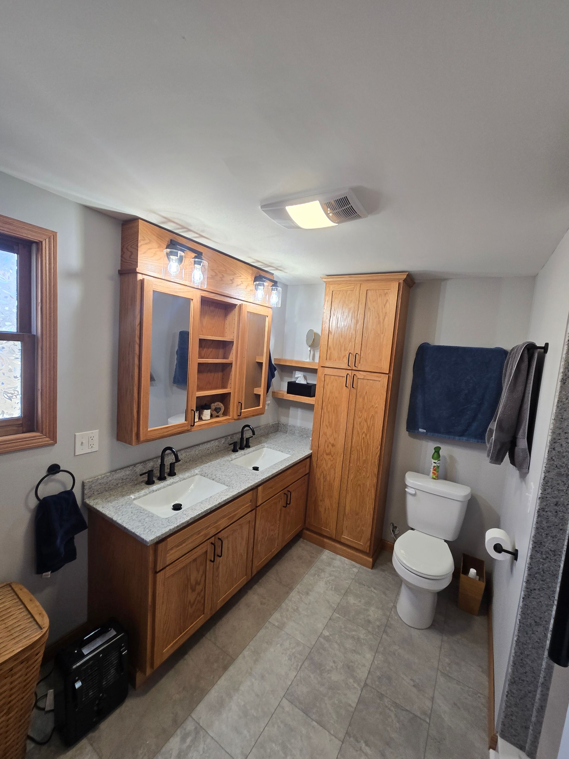 A bathroom with light-wood double vanity cabinets, a tall storage unit, light countertops, two sinks, and a toilet.
