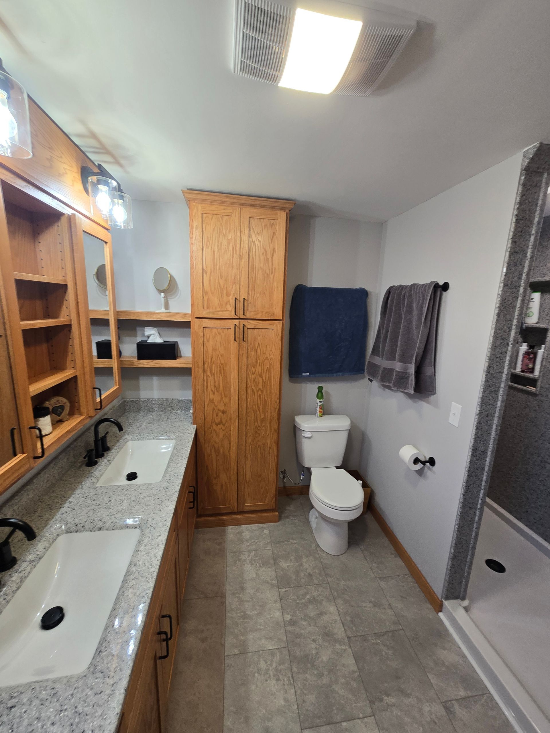 A modern bathroom with oak cabinetry, double sinks with granite countertops, a toilet, and a tiled walk-in shower.
