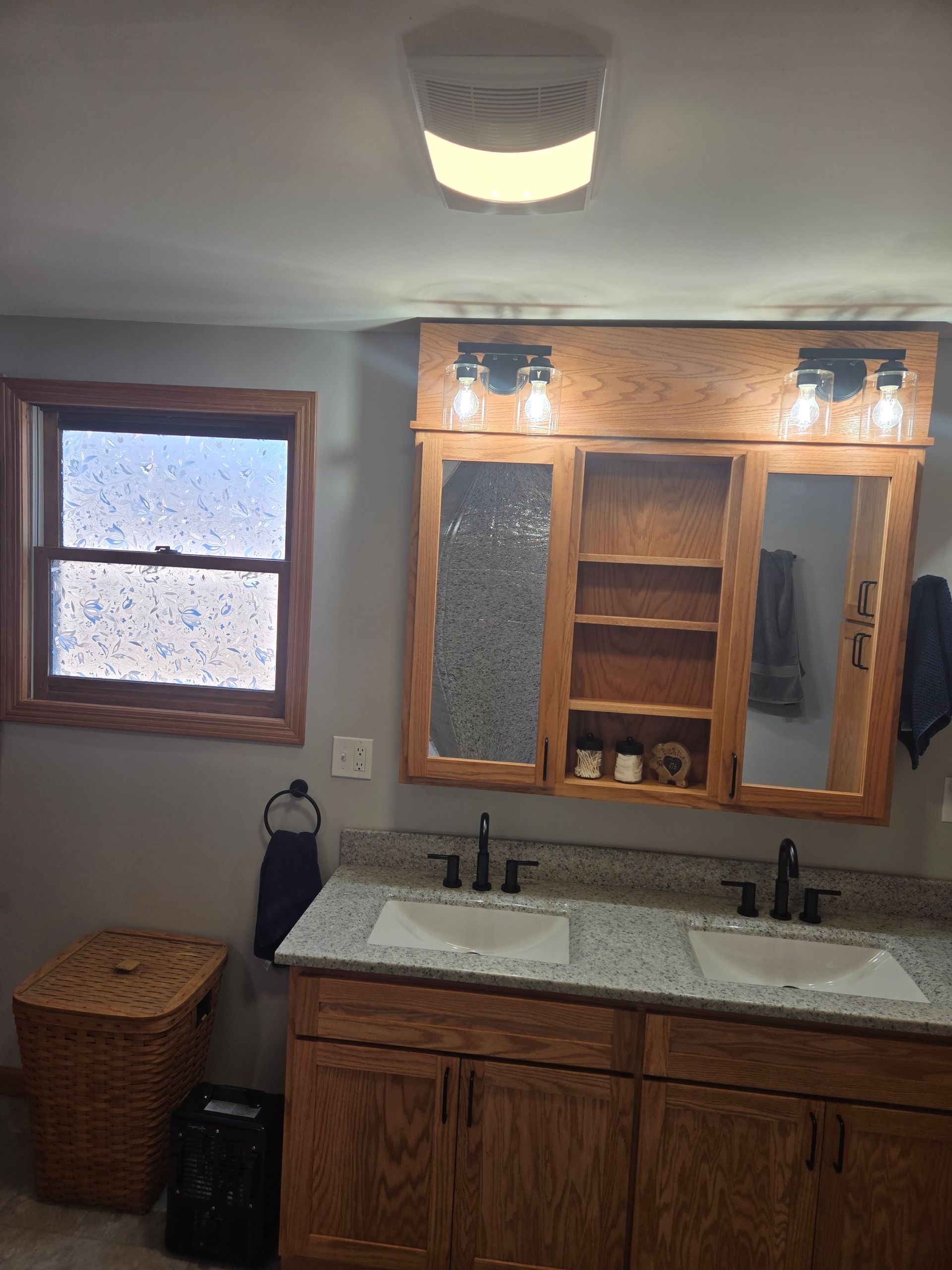 A bathroom vanity with wood cabinetry, a granite countertop with dual sinks, mirrored cabinets, and wall-mounted lights.