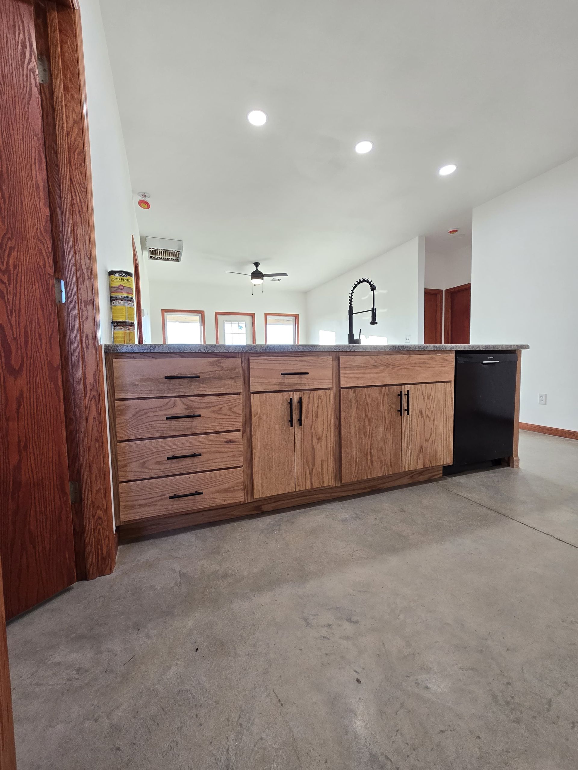 A kitchen island with wood cabinets, black hardware, and a black dishwasher set against a plain wall and concrete floor.