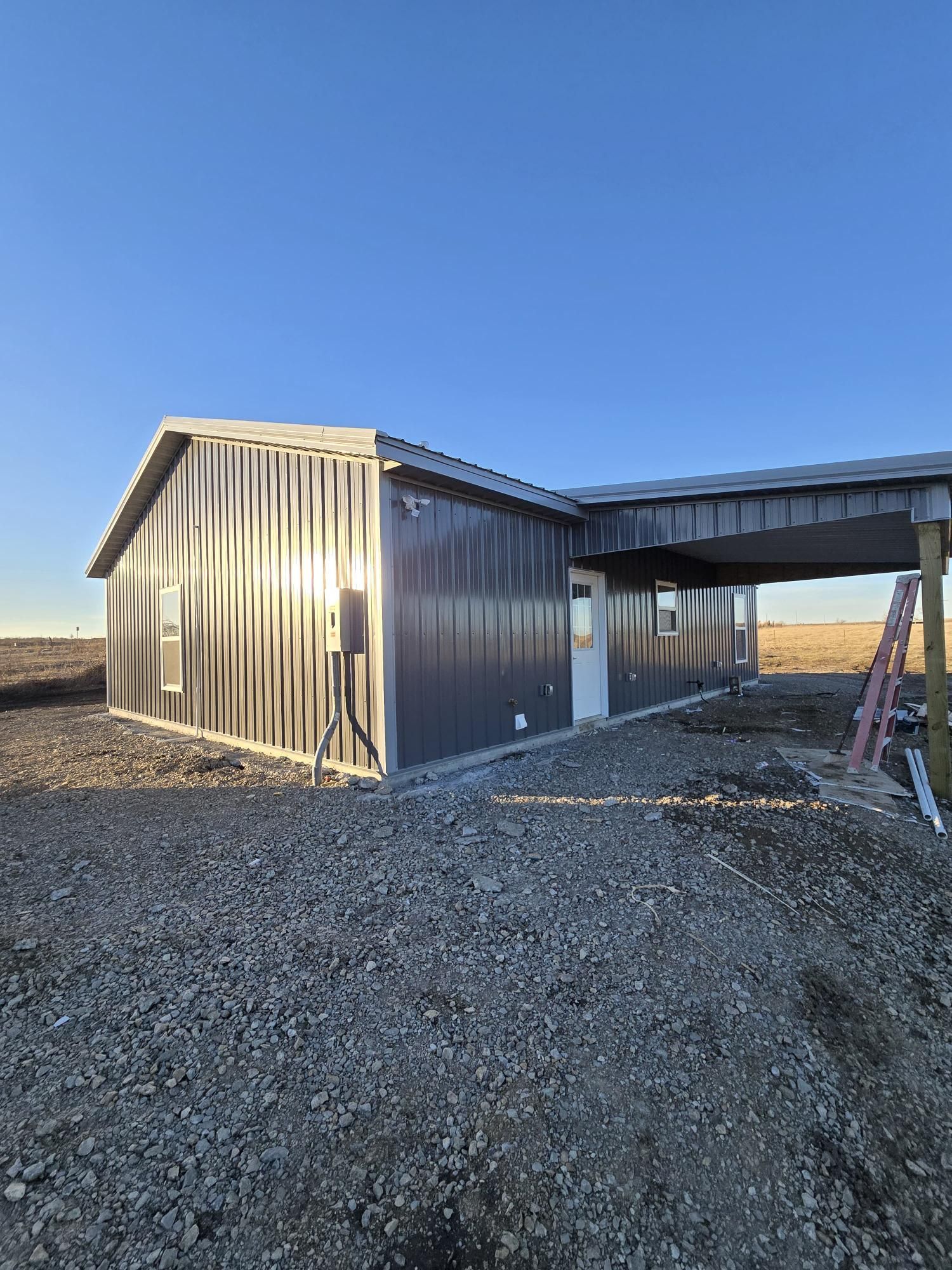 A side view of a gray metal-sided building under a blue sky, featuring a gravel yard and an attached open-sided porch.