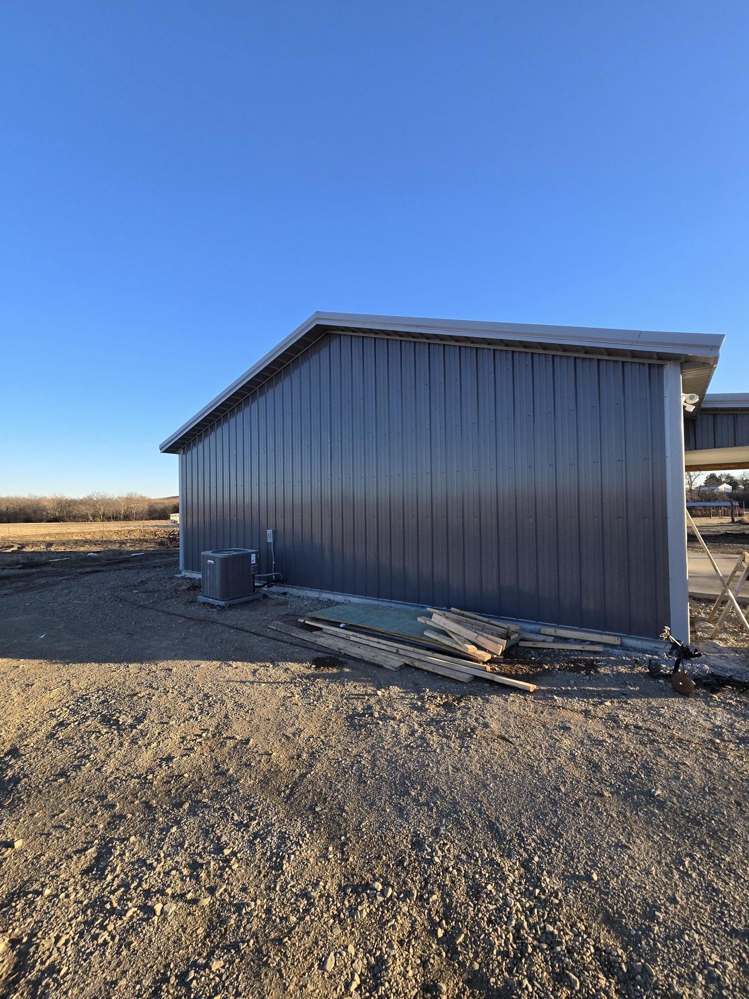 A grey metal-sided building stands on a gravel lot under a clear blue sky, with some construction debris at its base.