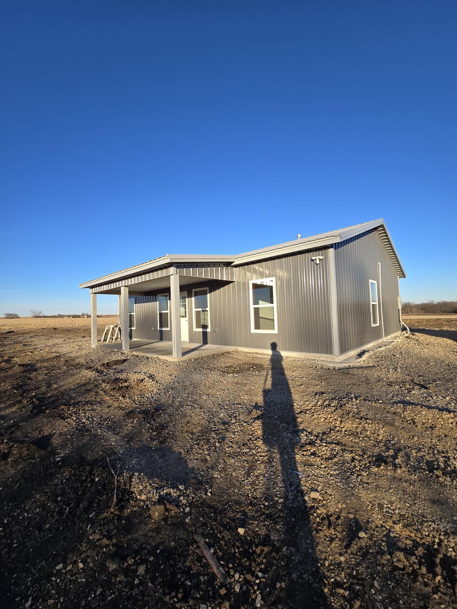 A grey, metal-sided barndominium-style house with a front porch sits on a dirt lot under a clear blue sky.