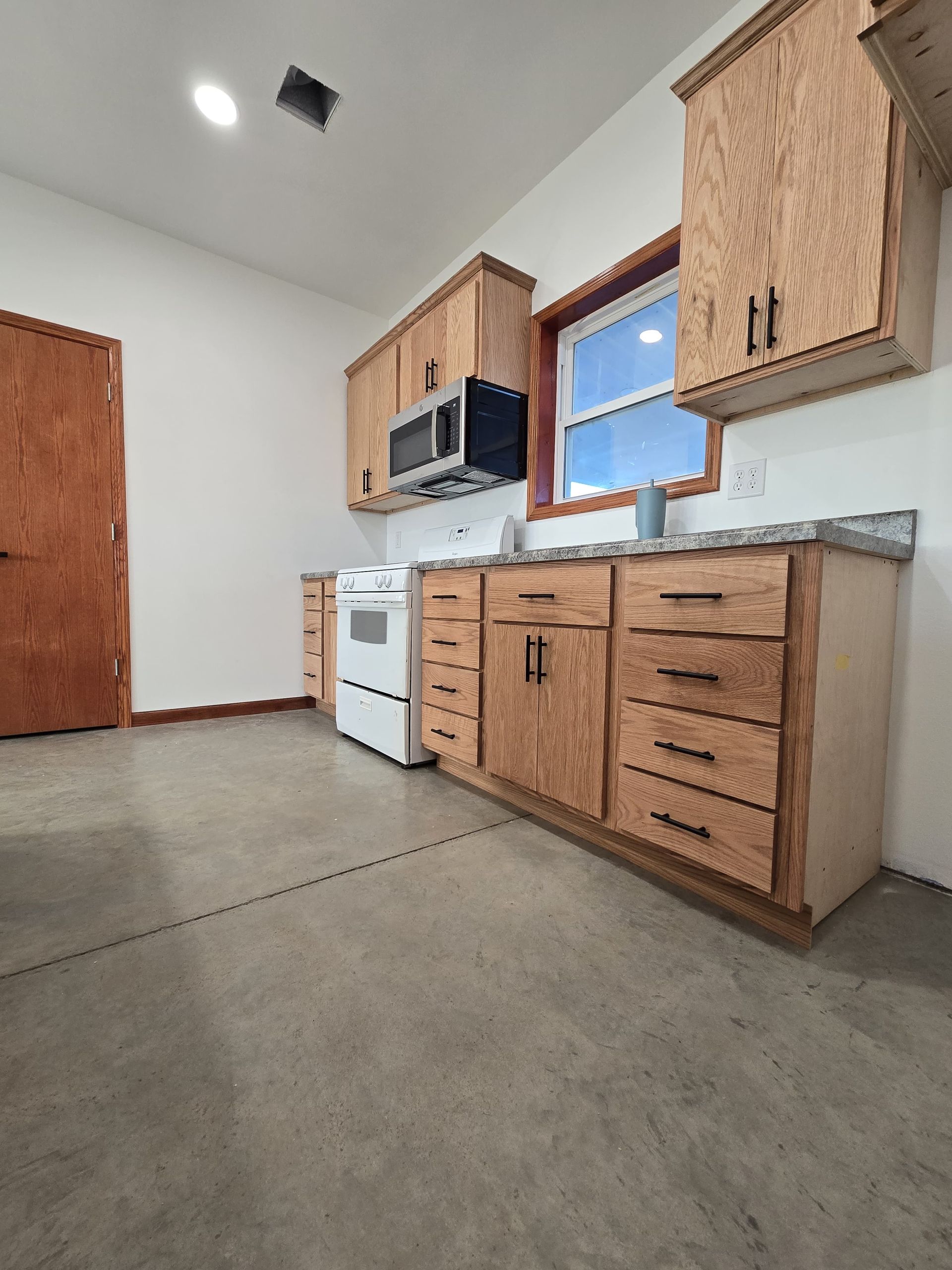 A small kitchen featuring natural wood cabinets, a white stove, gray countertops, and polished concrete floors.