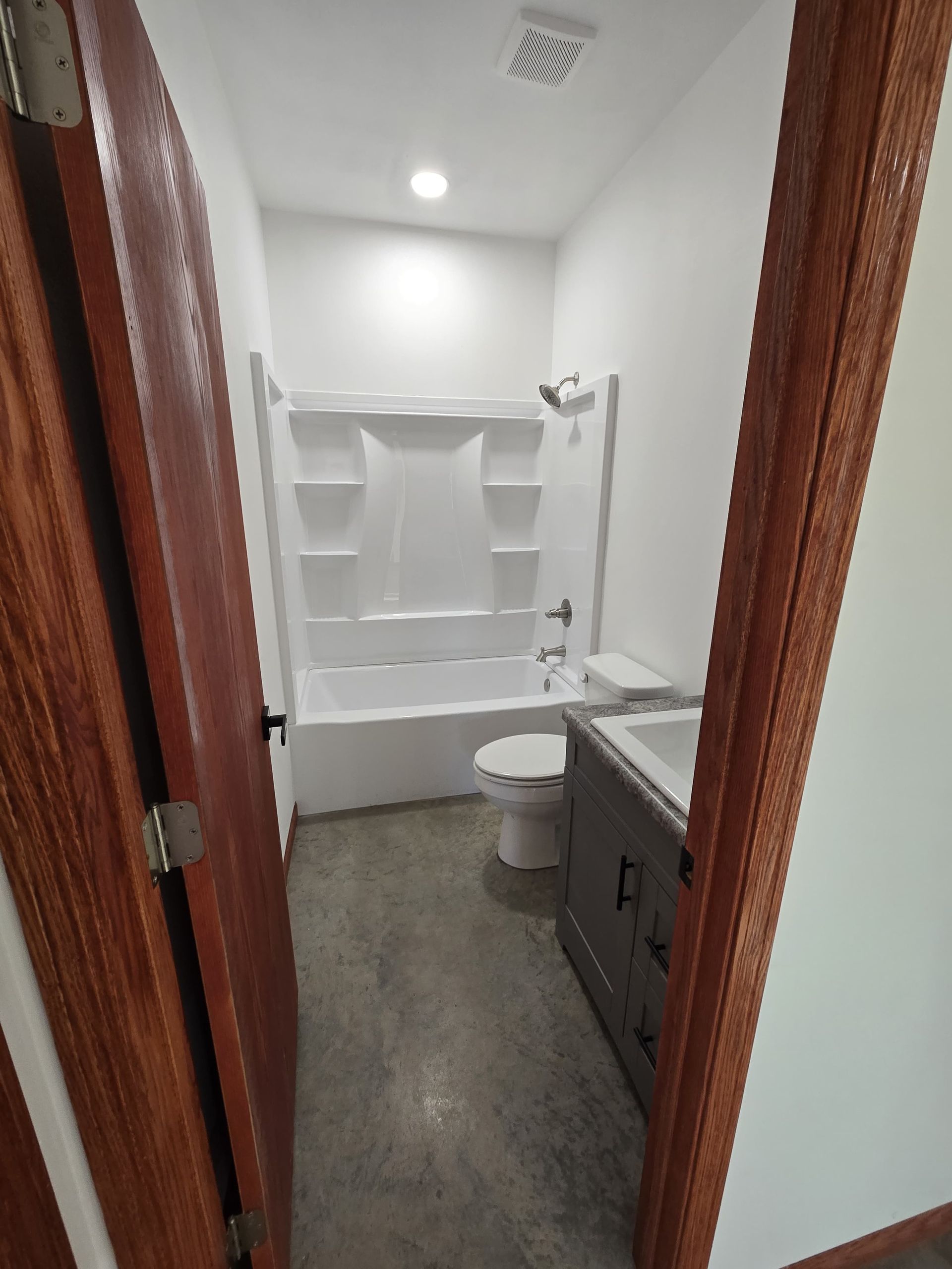 A narrow bathroom featuring a white tub-shower unit, a toilet, a grey vanity, and wood trim around the doorway.