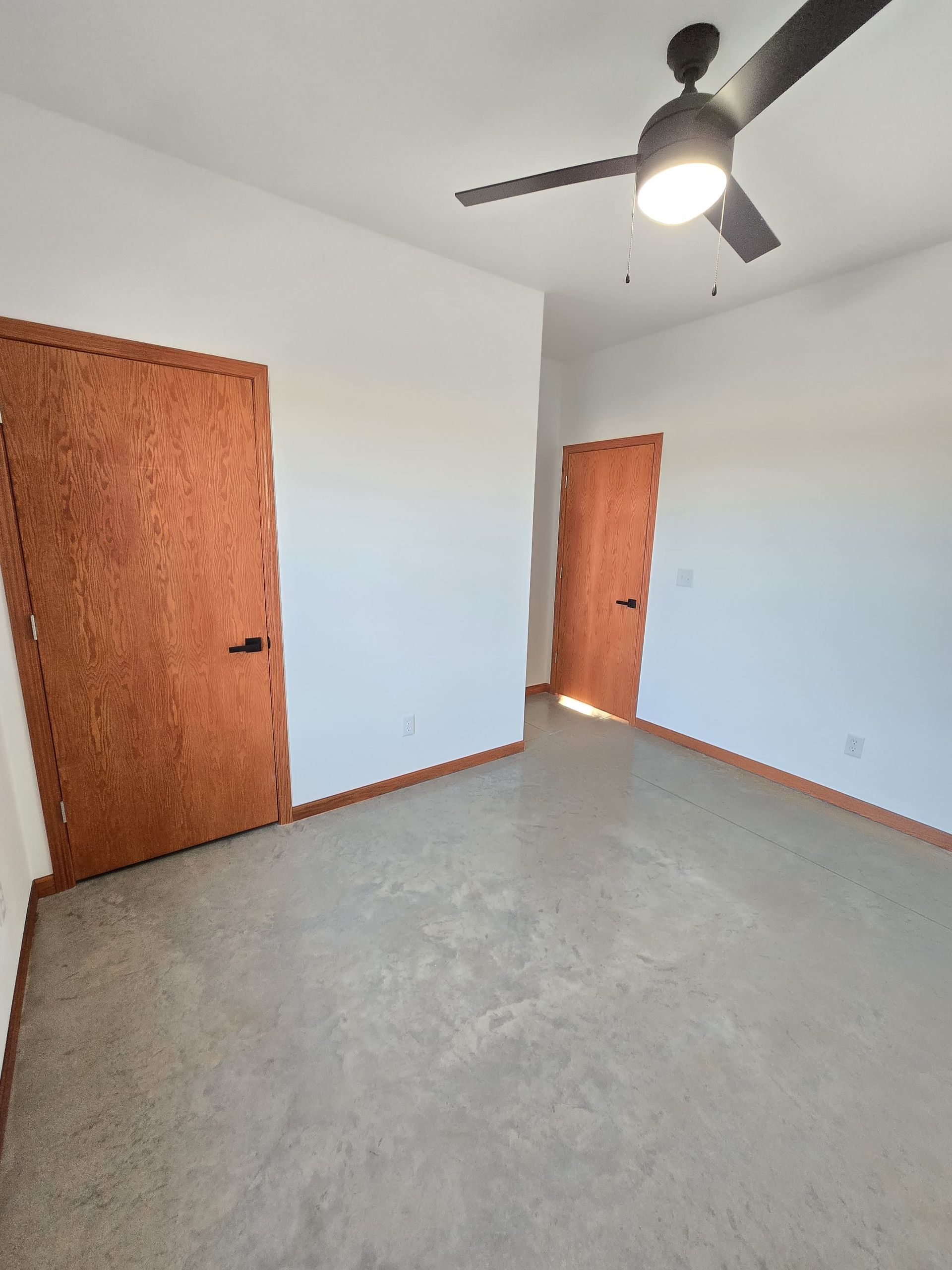 Empty room with white walls, two wooden doors, a light-colored speckled floor, and a ceiling fan.