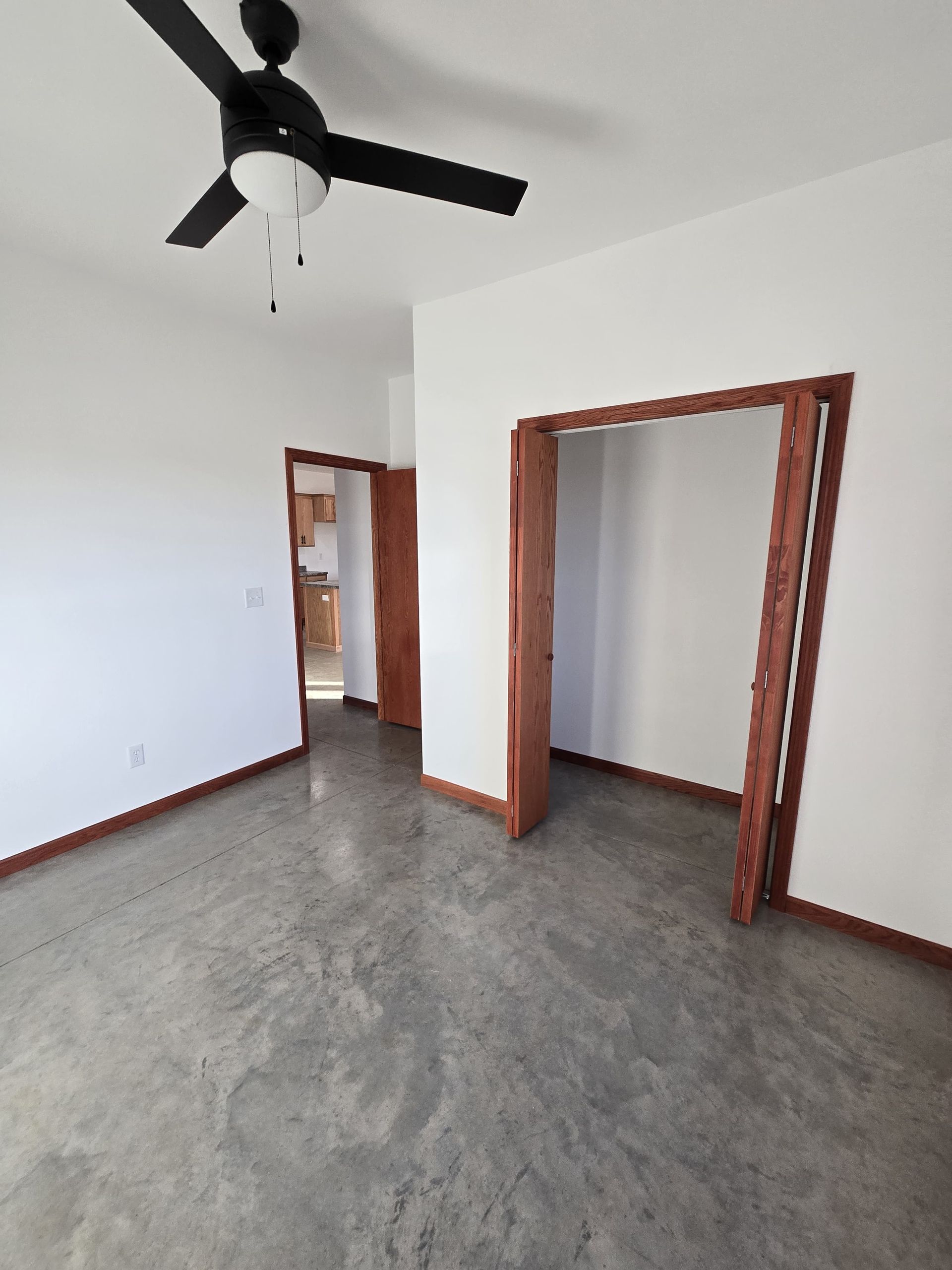 An empty room with gray polished concrete floors, white walls, a ceiling fan, and two open doorways with wood trim.