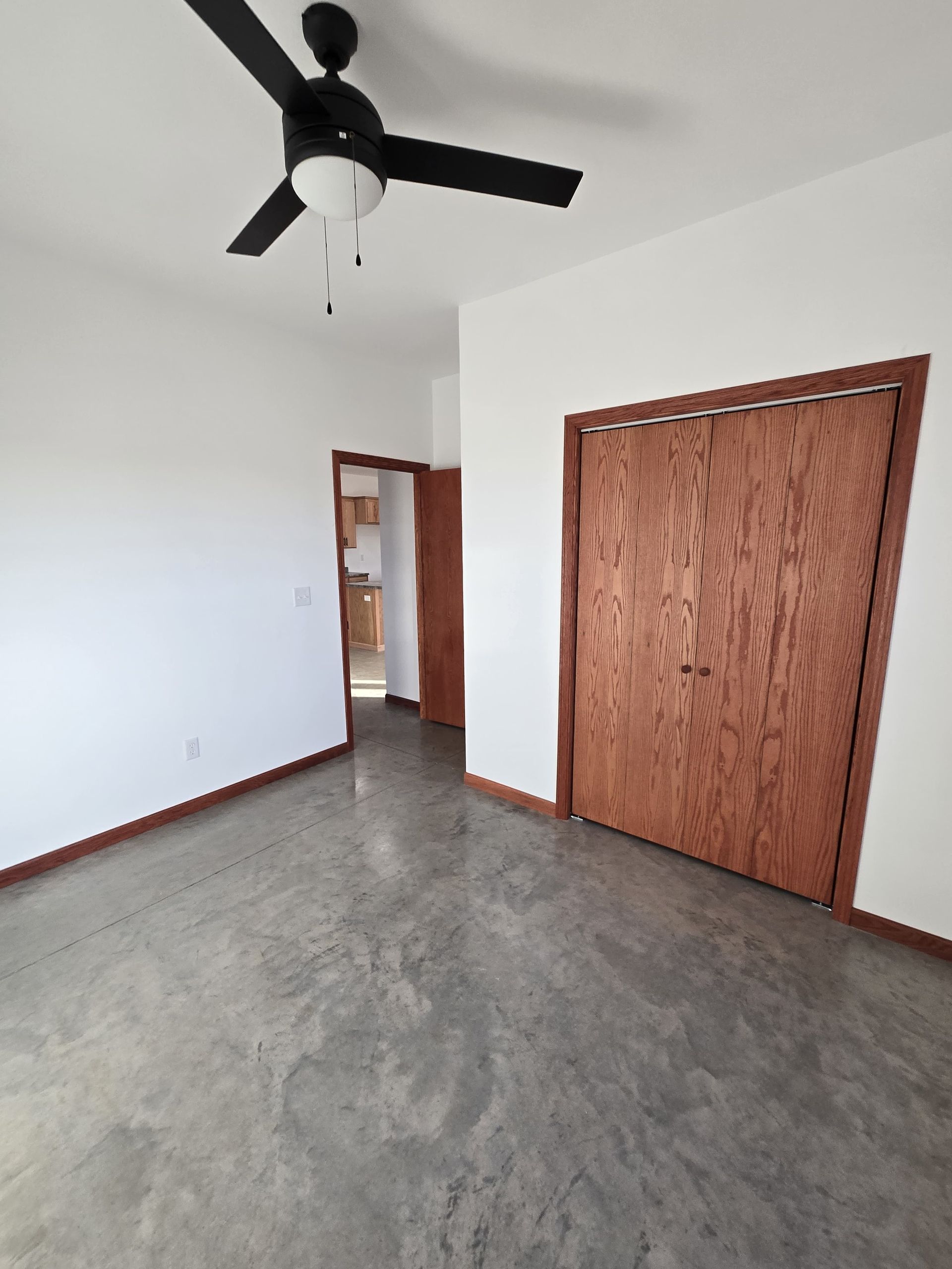 An empty bedroom with white walls, a black ceiling fan, wood-toned closet doors, and polished gray concrete flooring.