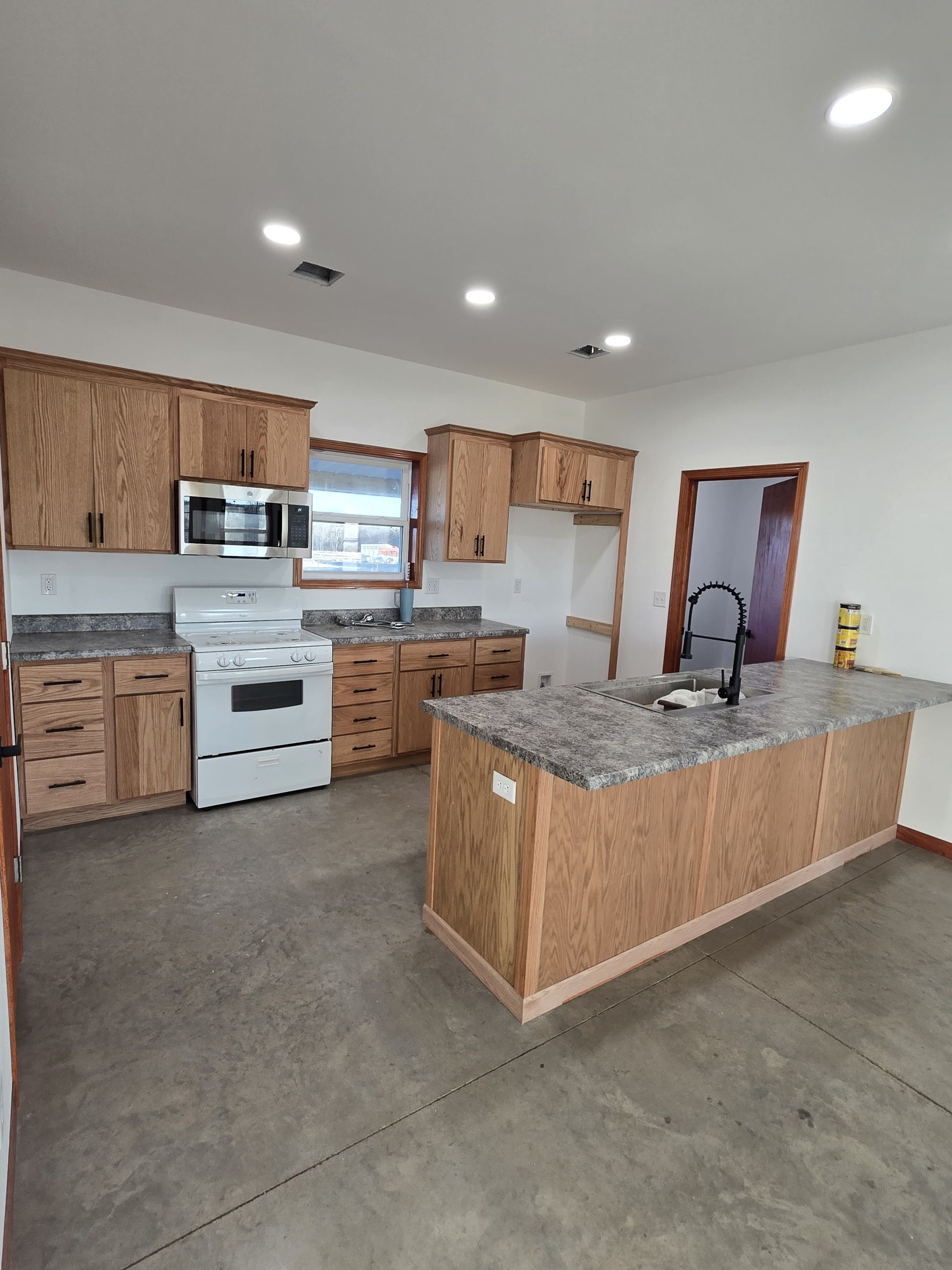 A modern kitchen with oak cabinets, granite countertops, white appliances, and a central island on a concrete floor.