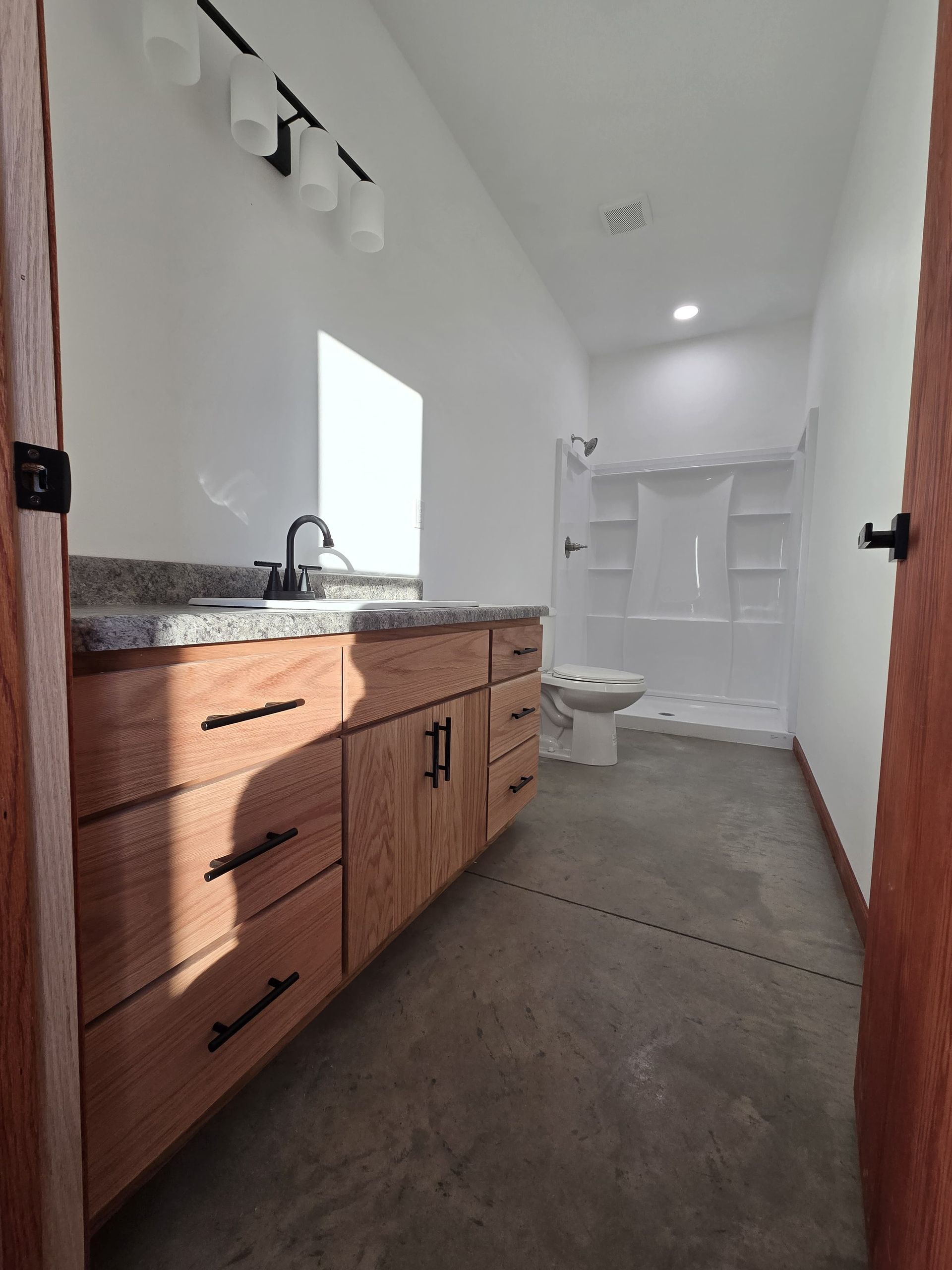 A modern bathroom with light wood cabinets, a granite countertop, concrete flooring, and a white shower stall.