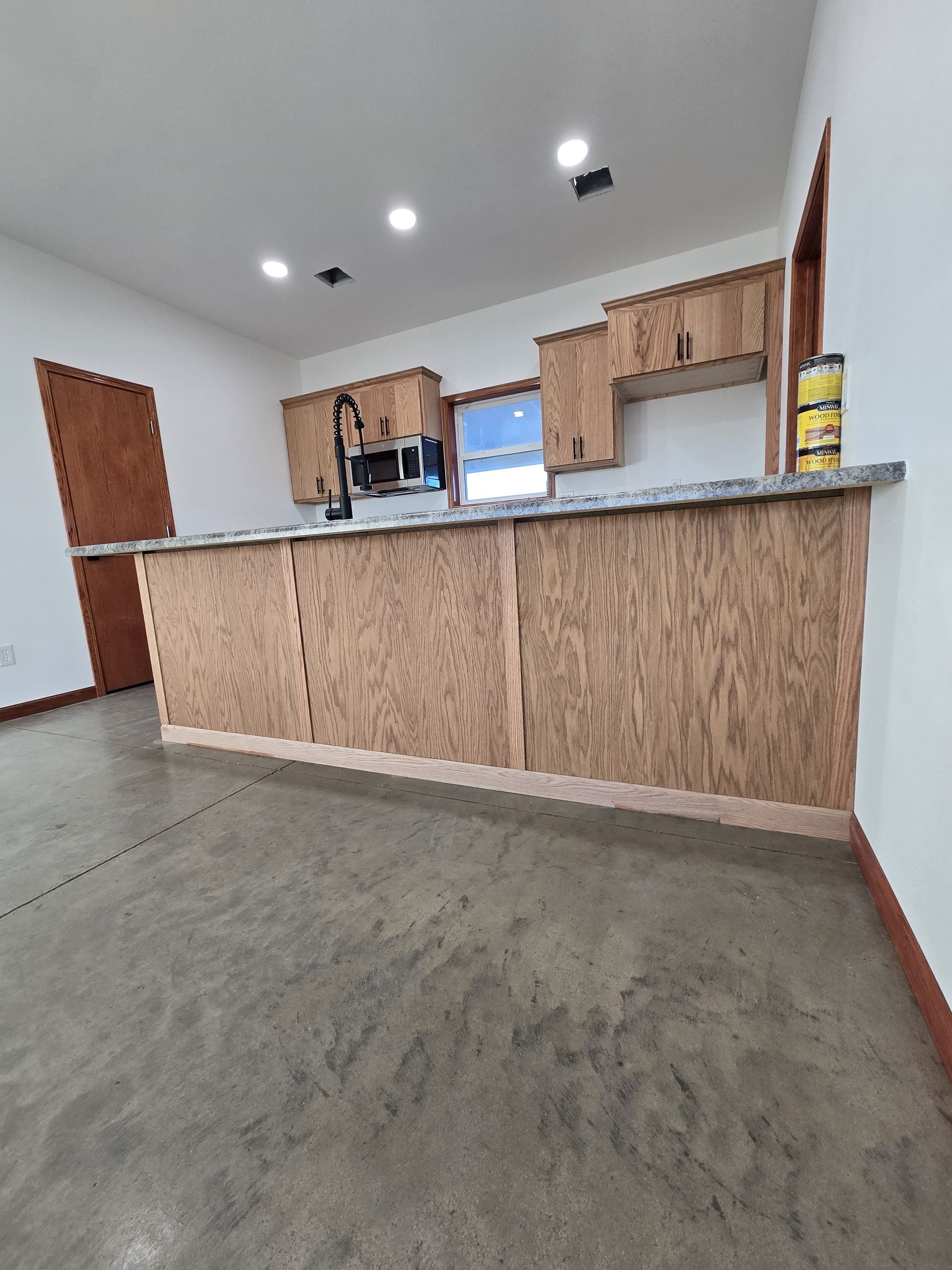 A kitchen island with unfinished wooden panels, topped with granite, in a room with white walls and a grey concrete floor.
