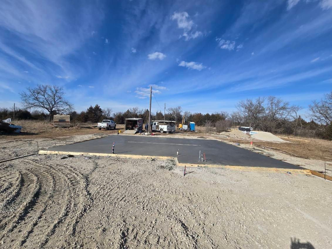 A rectangular concrete foundation slab sits on a gravel lot under a sunny blue sky with thin clouds.