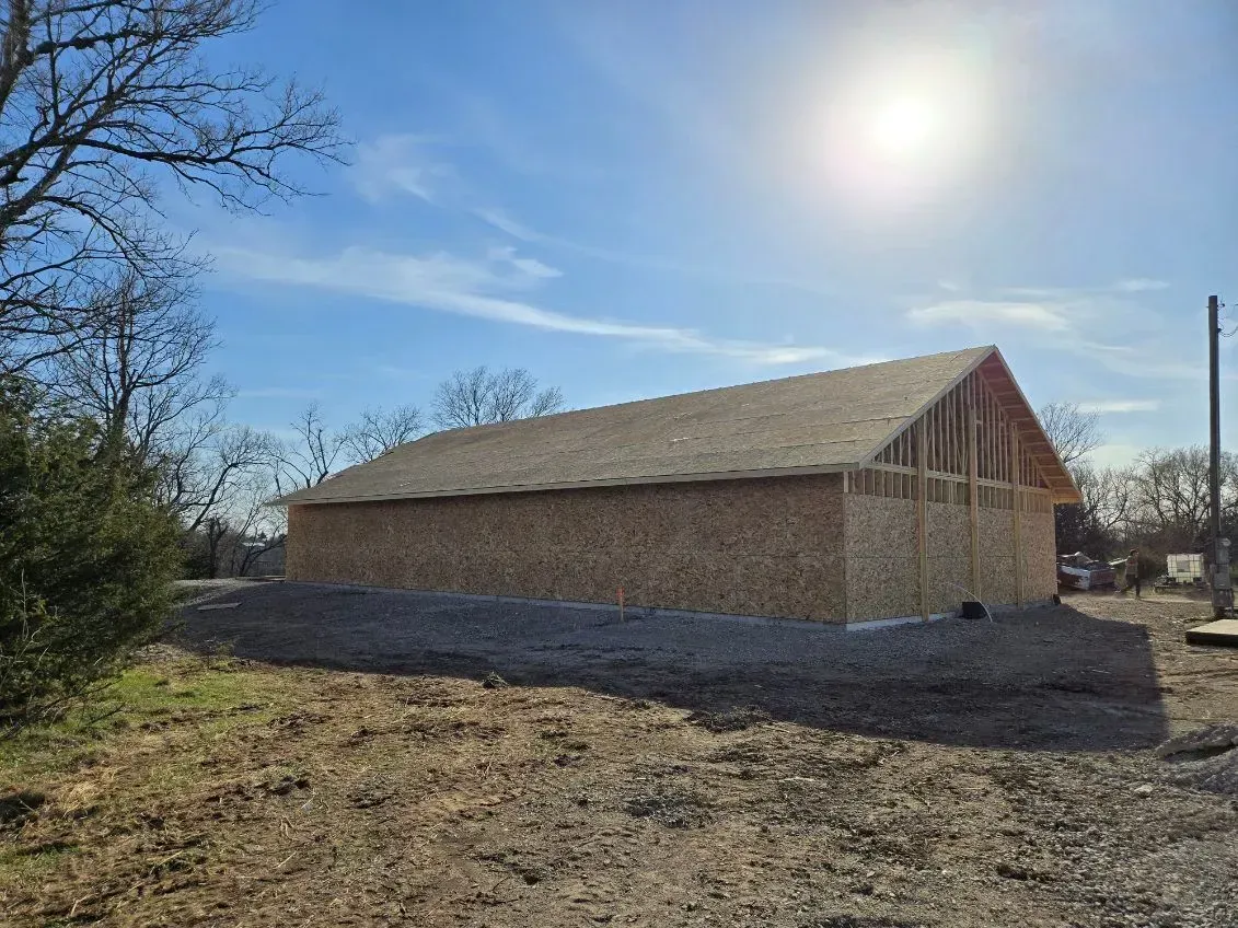Tan metal shed with a brown roof and trim, set on a gravel base against a backdrop of green trees and a blue sky.