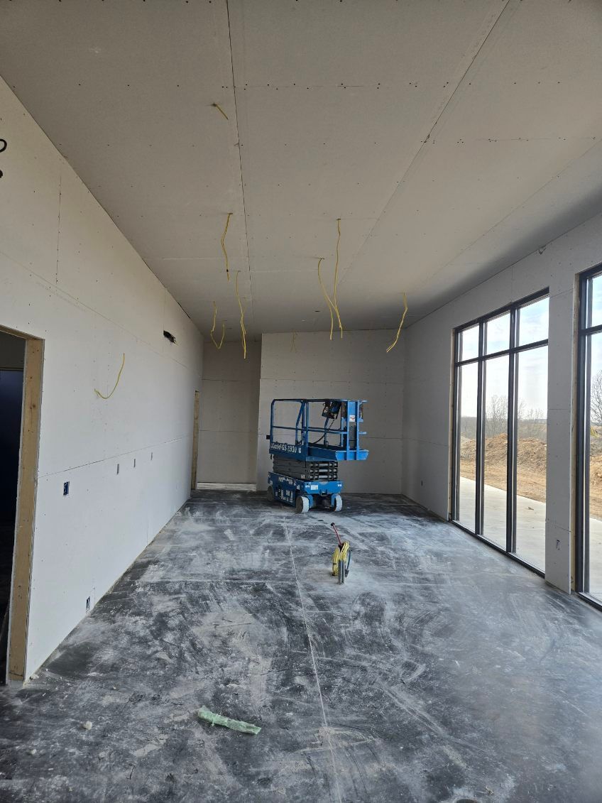 An unfinished room with drywall on walls and ceiling, a blue scissor lift in the back, and large windows on the right.