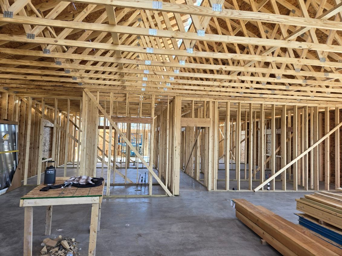 Framed house interior under construction with exposed wooden trusses, studs, and stacked lumber on concrete floor