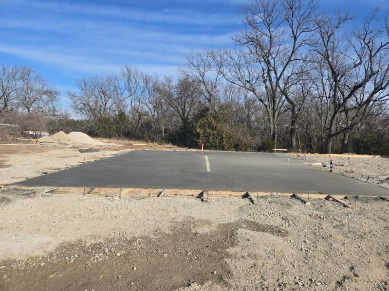 A freshly poured concrete slab foundation sits in a dirt clearing against a backdrop of bare trees under a blue sky.