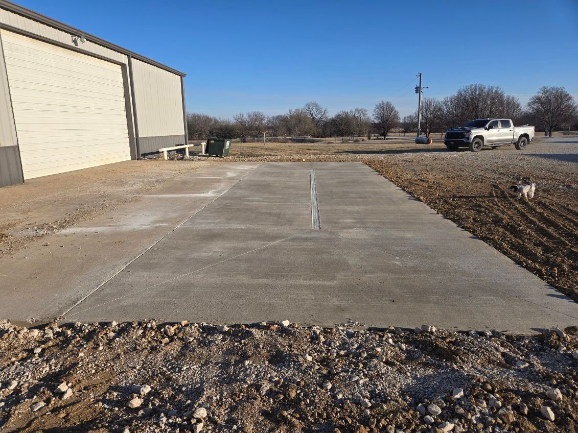 A rectangular concrete slab sits on gravel next to a metal building under a clear blue sky, with a truck parked nearby.