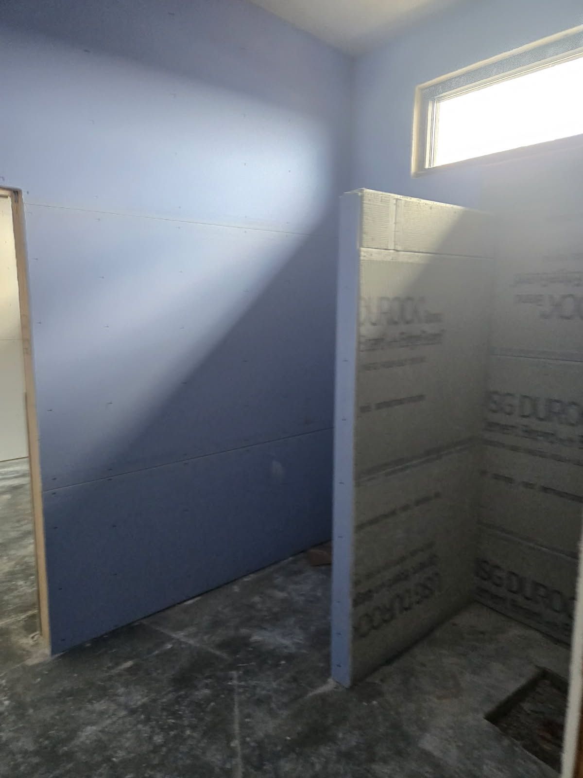 An unfinished room with blue-painted walls and a shower partition covered in grey cement board under a small window.