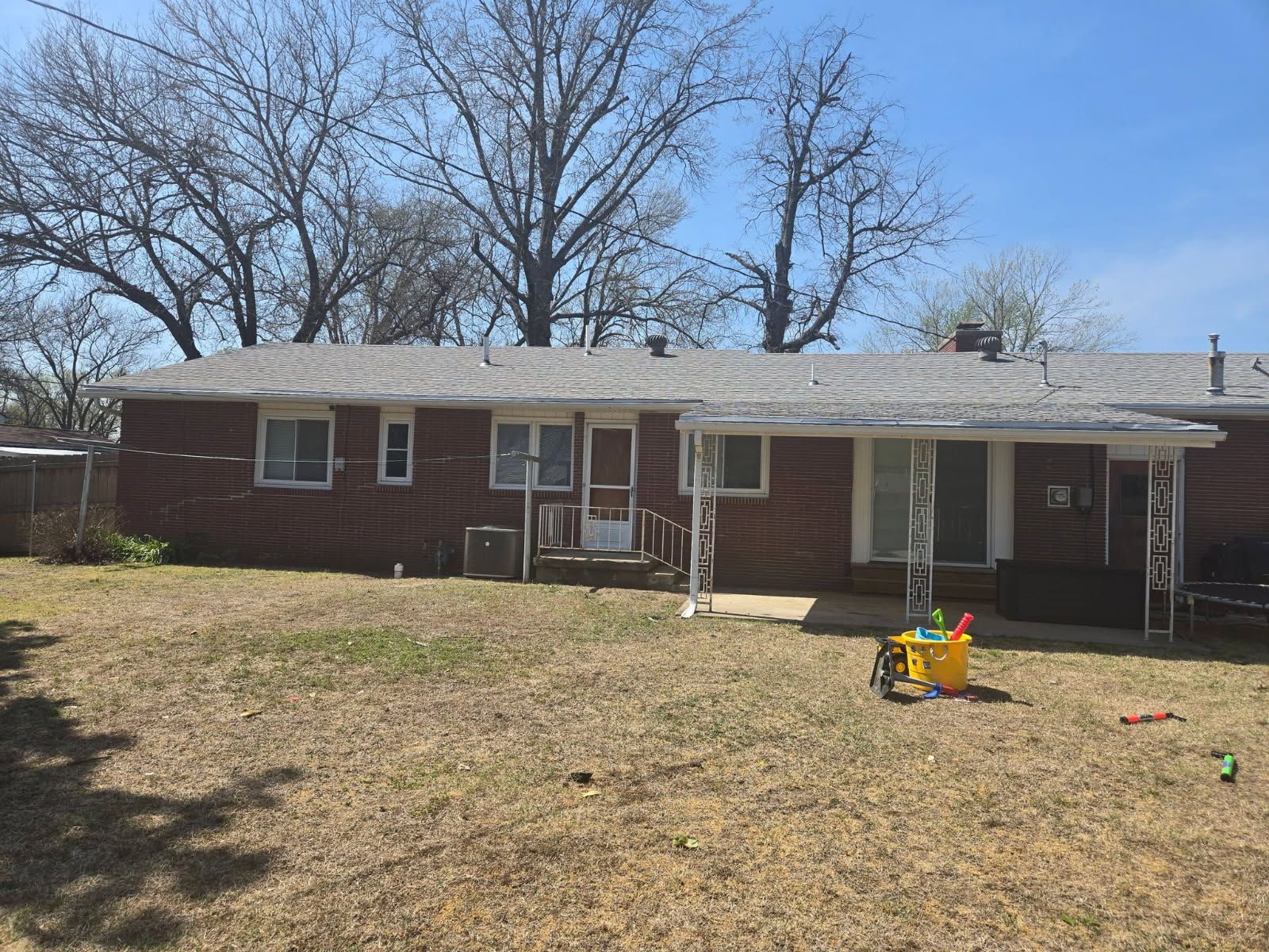 Backyard view of a brick house with a covered patio, bare trees, and a children’s toy on the lawn