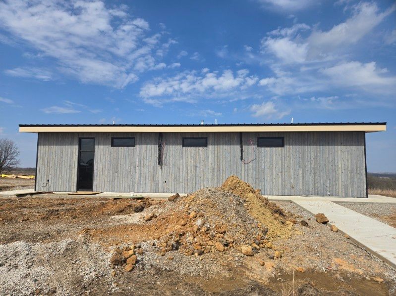 A single-story building with gray wooden siding, a black door, three small windows, and a pile of dirt in the foreground.