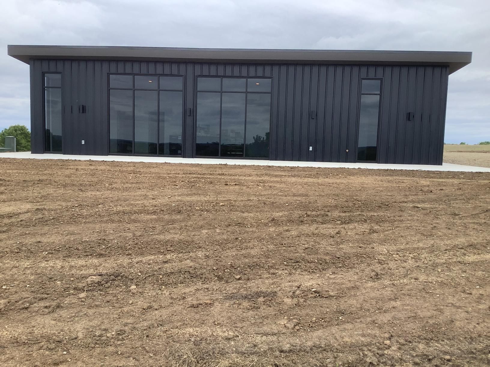 Modern black commercial building with large windows, set behind a bare dirt foreground under a cloudy sky