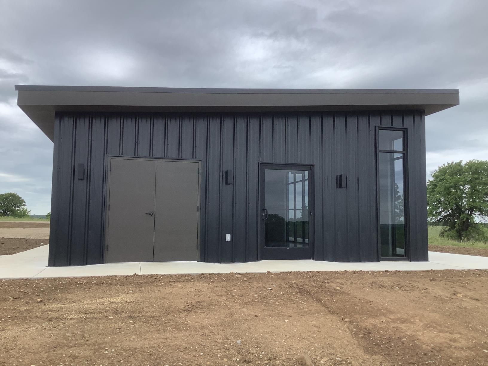 Small black modern utility building with a gray door and glass entrance under an overcast sky