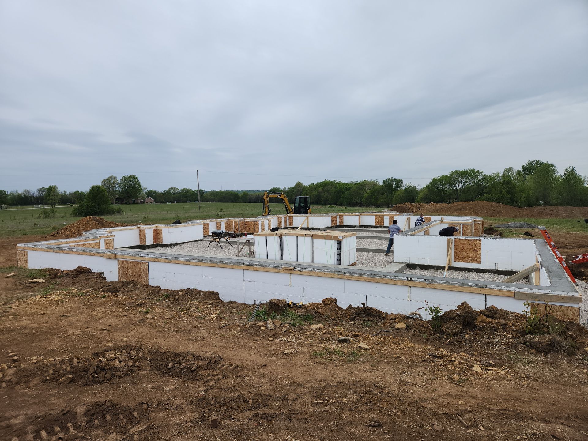 Foundation of a house under construction with white foam blocks and wood supports on a dirt lot under an overcast sky.