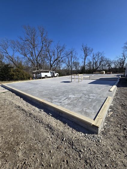 A concrete building foundation with wooden framing boards on the edges, set in a dirt lot under a clear blue sky.