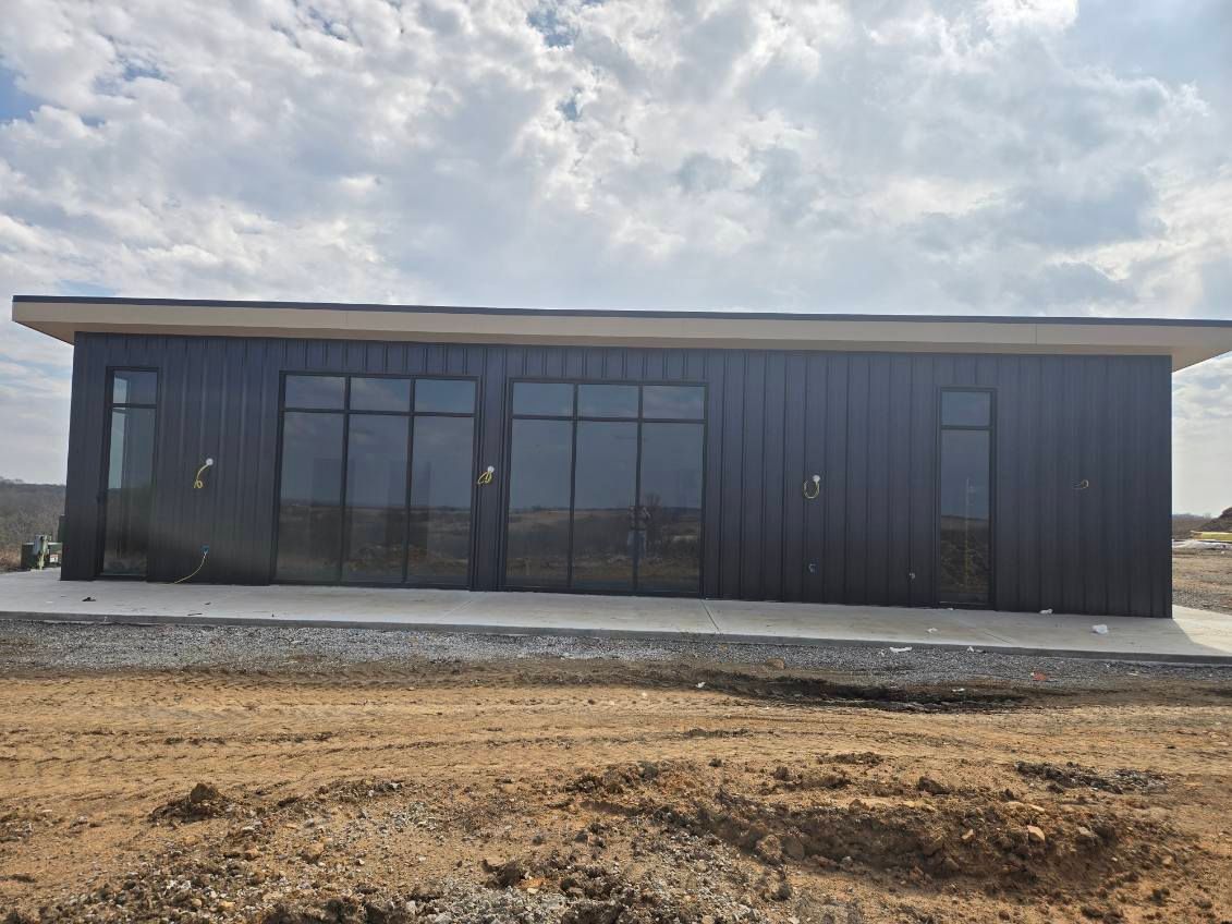 A modern, dark-sided rectangular building with large glass doors and windows, set against a cloudy sky and dirt ground.
