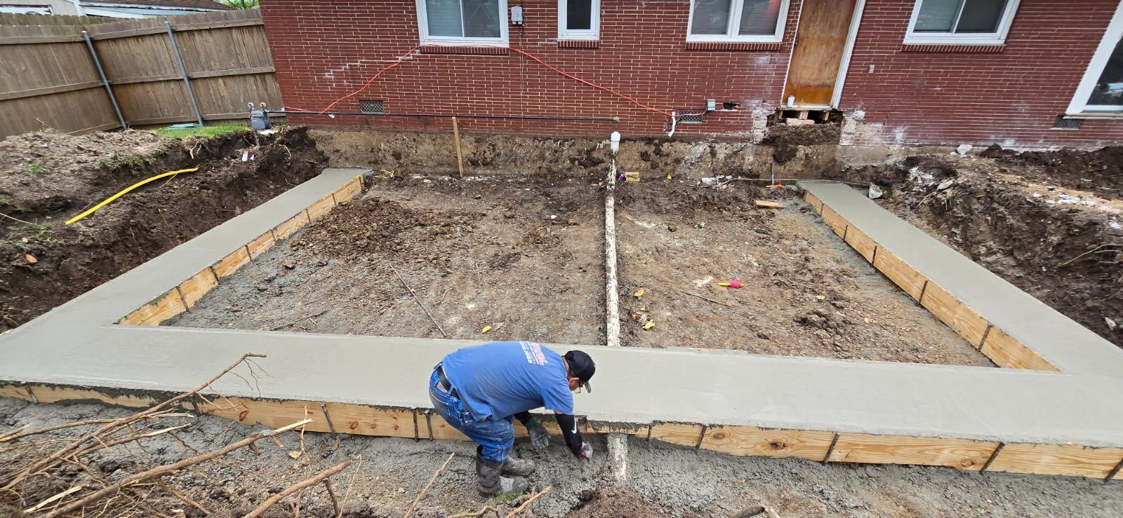 Worker kneeling at a concrete foundation frame beside a brick house, with bare ground inside the lot.