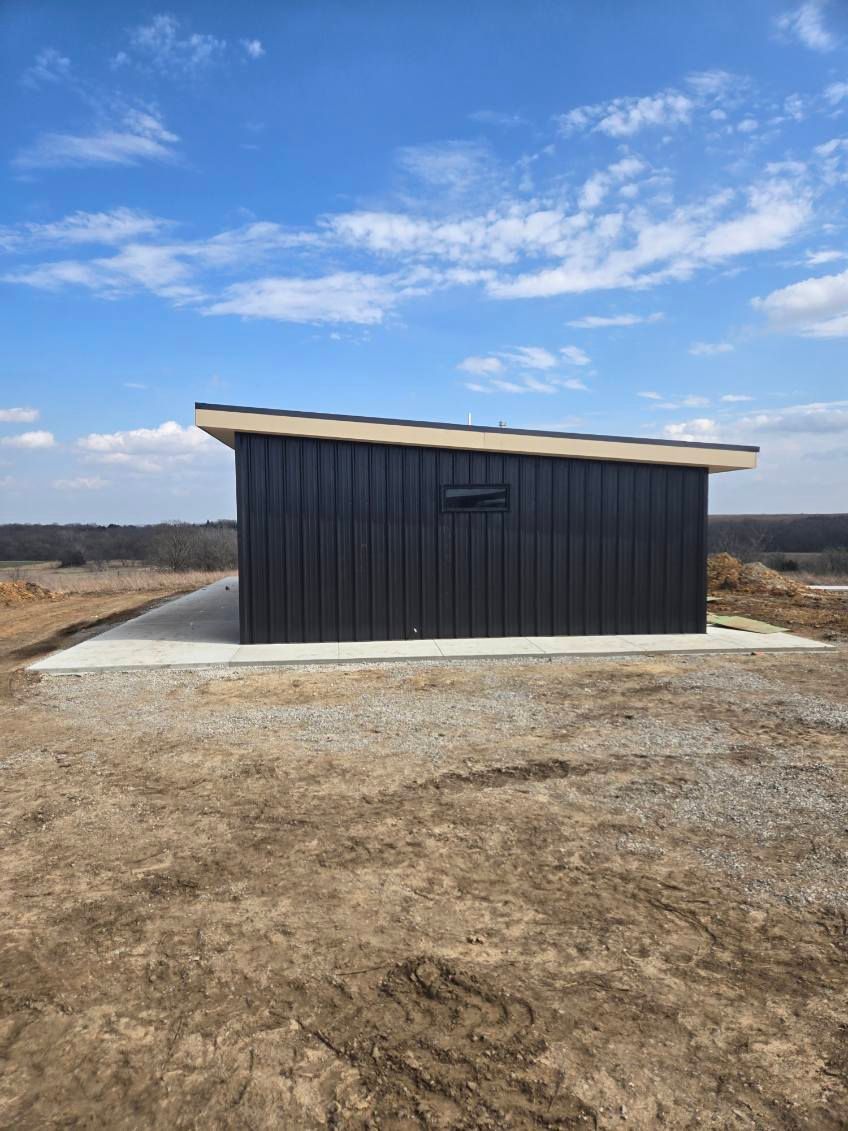 A modern black shed with a slanted roof sits on a concrete pad in a rural, open landscape under a sunny, cloudy sky.