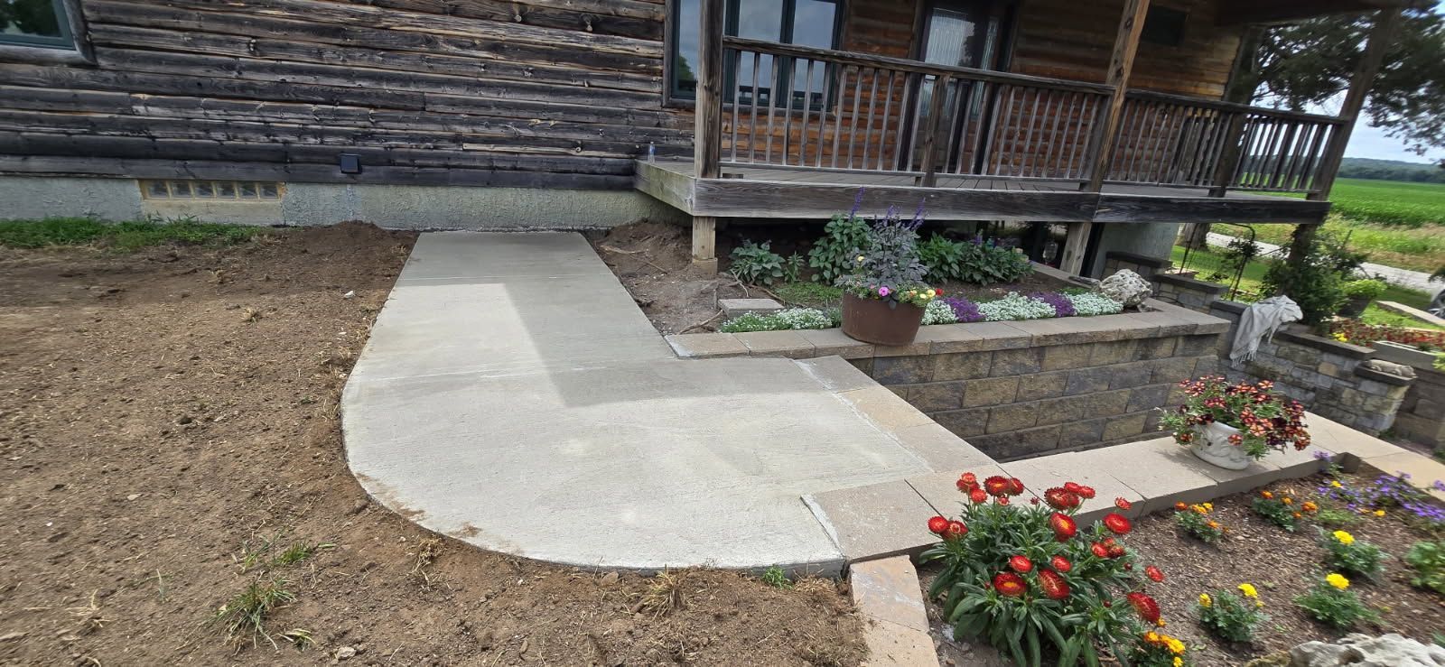 Concrete walkway in a garden beside a wooden house, with flower beds and potted plants.