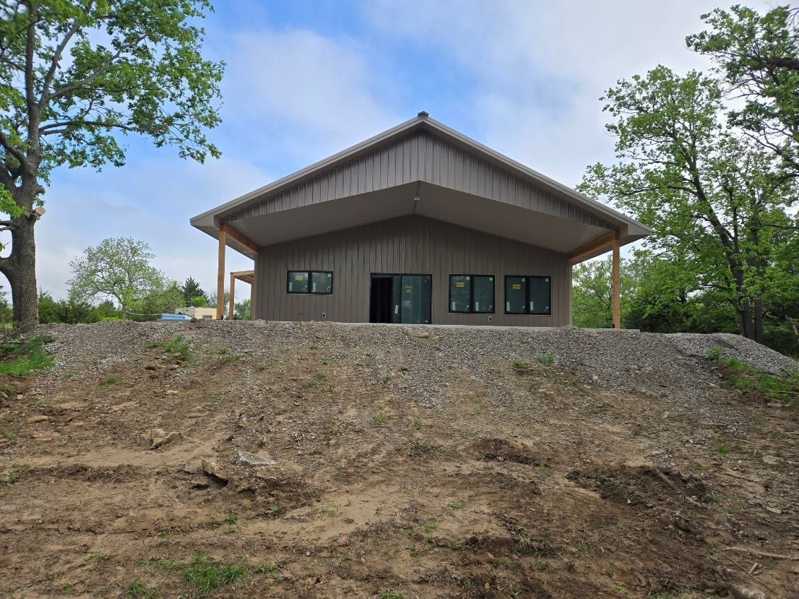 Modern cabin-style building on a wooded hillside, with a steep roof and gravel embankment in front