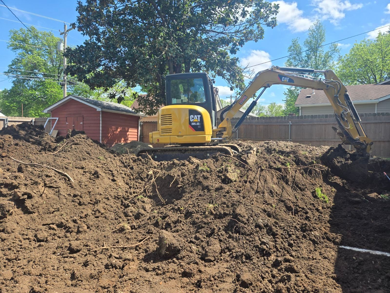 Yellow excavator digging a large dirt pile beside a red shed and fence under a blue sky