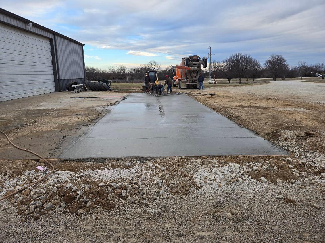 A concrete truck pours fresh cement for a new driveway section next to a metal building in an outdoor setting.