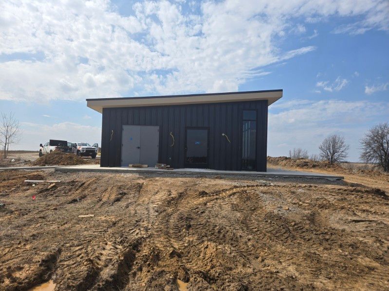 Tan metal barn with brown trim and an open front entrance, a side overhang, and gravel ground.