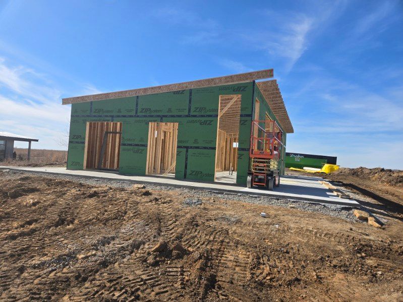 A new home under construction with green sheathing, wooden framing, and a concrete foundation on a sunny, dirt lot.