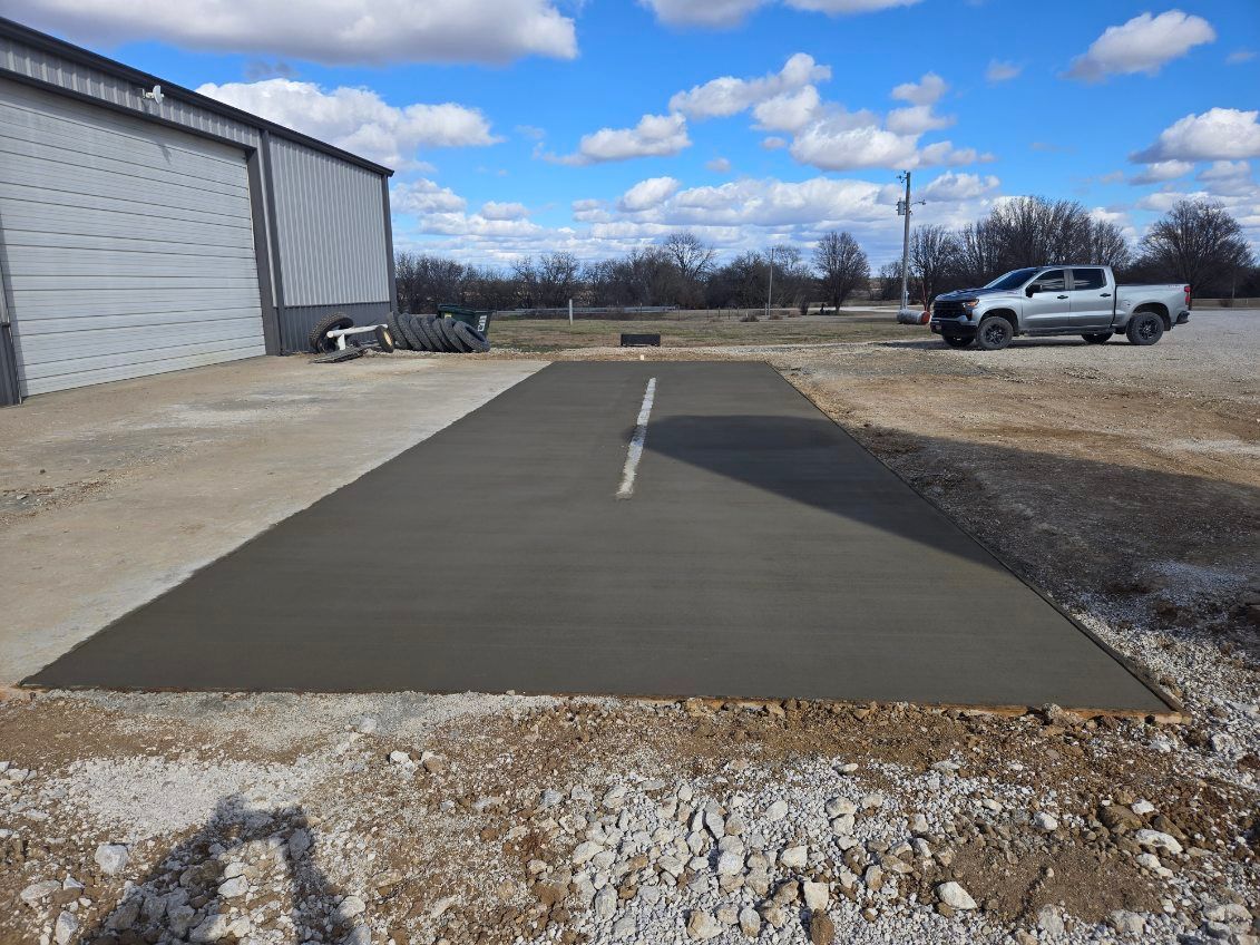 A freshly poured rectangular concrete pad sits on a gravel lot next to a metal building and a parked pickup truck.