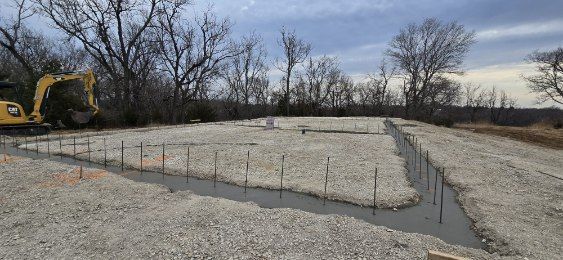 A yellow excavator prepares a foundation site with a gravel base and water-filled trenches for a building project.