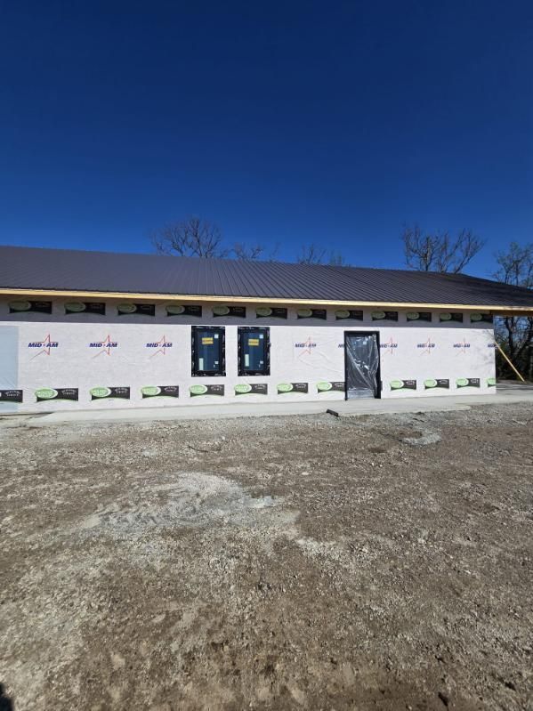 Single-story house under construction with wrapped exterior walls and gravel foreground under a clear blue sky