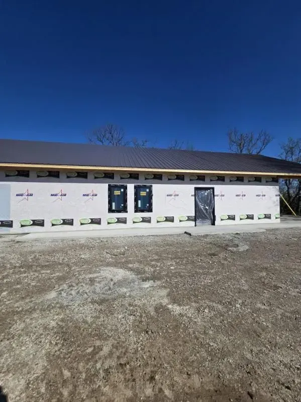 Tan metal building with a brown trim and gravel path, next to a dirt path with trees on the left. Blue sky in the background.