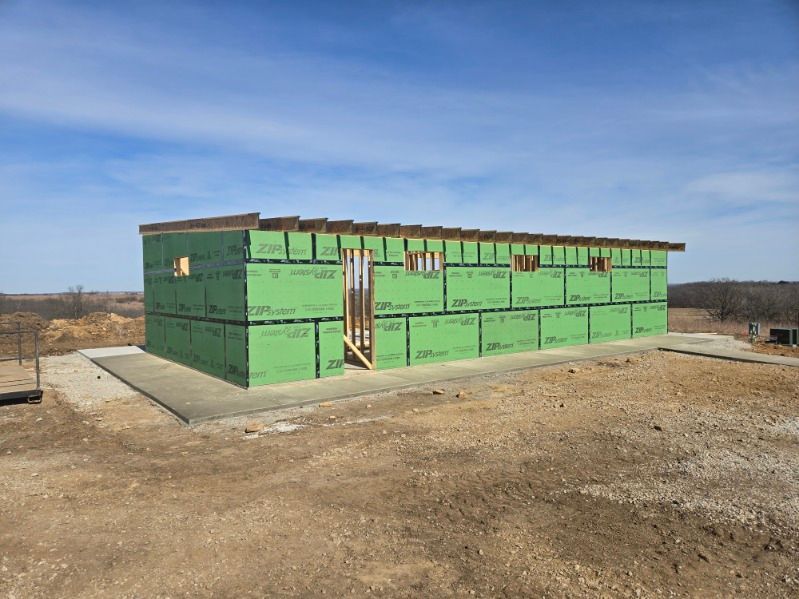 A rectangular building frame under construction, covered in green wall sheathing, set on a concrete slab in an open field.