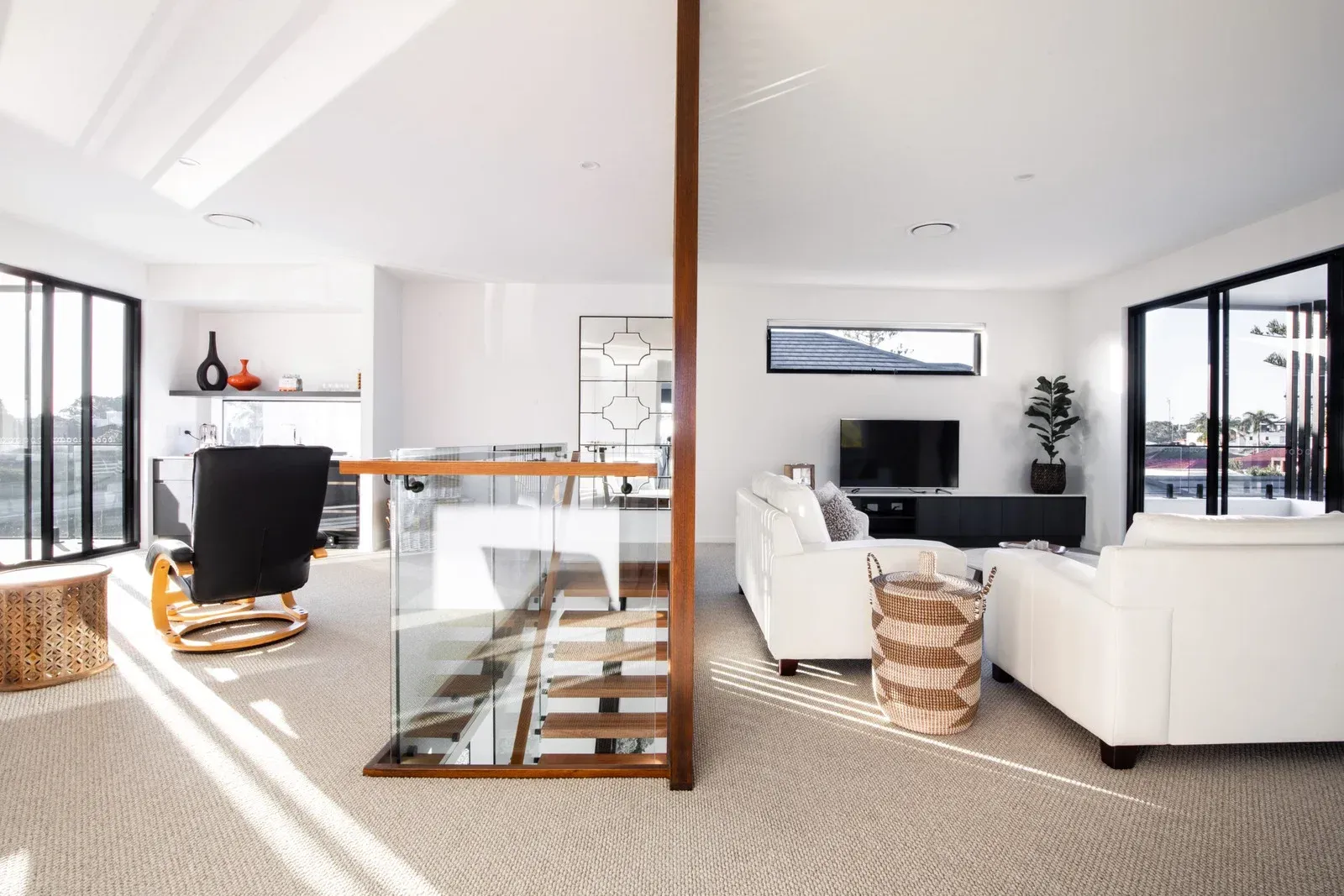 Modern living room with stairs, glass railings, white walls, and dark wood accents.