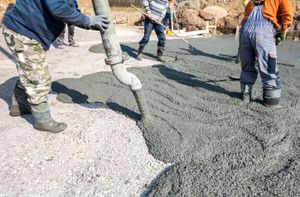 Workers pouring concrete onto a gravel base. A hose dispenses gray concrete; other workers use tools to spread it.