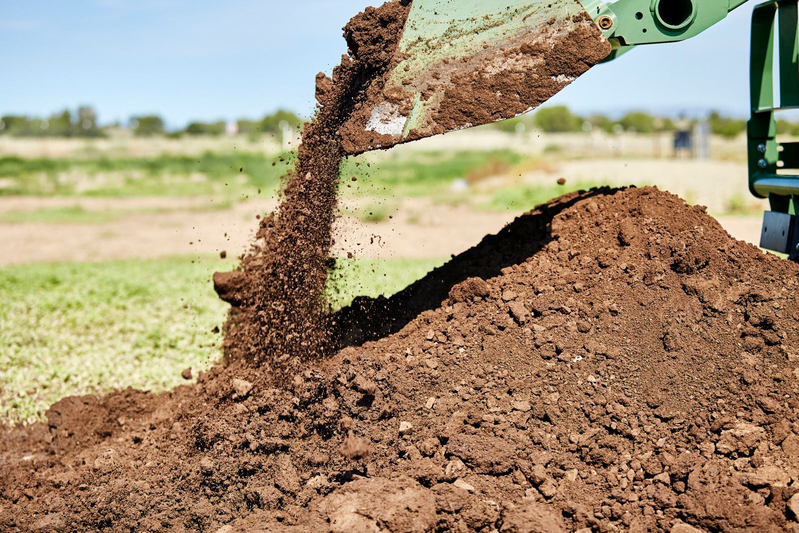 A backhoe dumping a pile of brown dirt onto a grassy field under a clear blue sky.