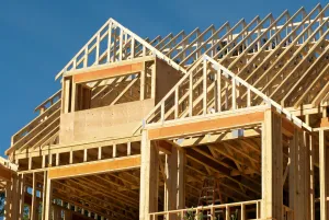 Wooden frame of a house under construction against a clear blue sky. Sunlight illuminates the exposed beams.