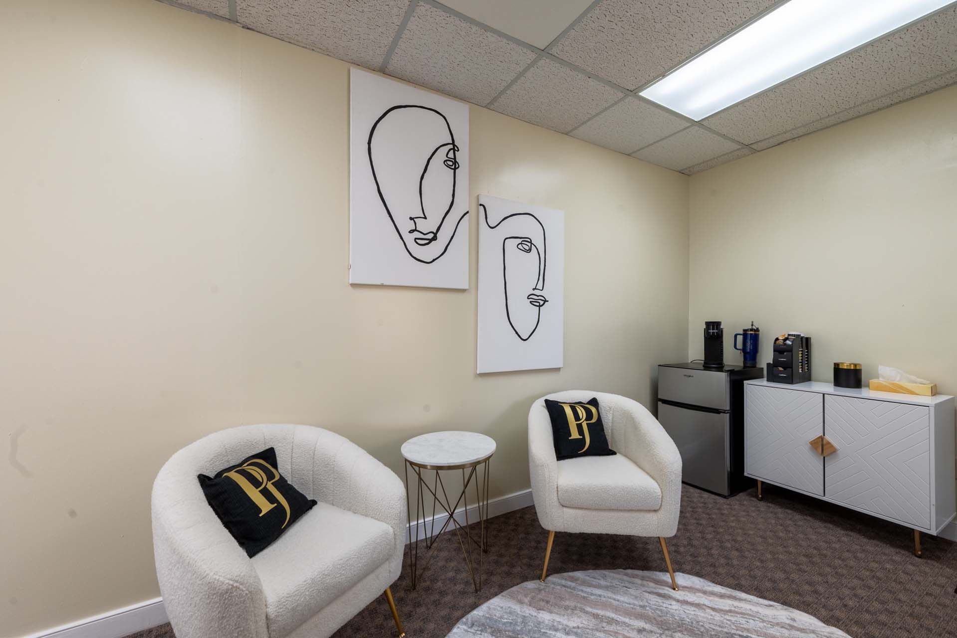 Two white armchairs in a beige room with abstract art, coffee station, and small side table.