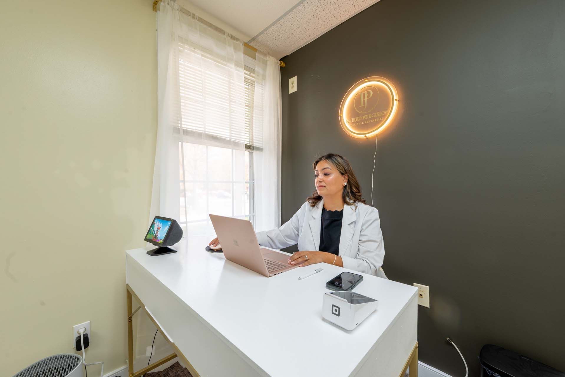 Woman at a white desk in an office, working on a laptop. There is a window and a neon sign.